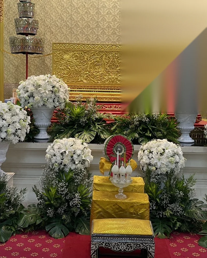 พุ่มลิลลี่และกุหลาบขาวในแจกันขาวหลายจุด ใบไม้เขียวเข้มรอบฐาน ฉากสลักลายทอง ฉัตรด้านซ้าย พรมแดง